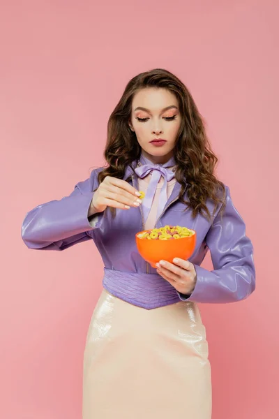Concepto de fotografía, mujer morena con el pelo ondulado fingiendo ser una muñeca, sosteniendo tazón con hojuelas de maíz, mirando el desayuno, posando sobre fondo rosa, chaqueta púrpura de moda - foto de stock