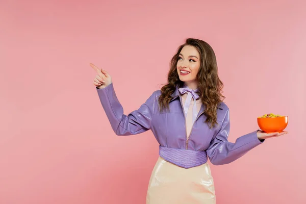 Concept photography, cheerful woman with wavy hair pretending to be a doll, pointing away, holding bowl with corn flakes, eating tasty breakfast, posing on pink background, stylish purple jacket — Stock Photo