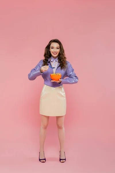 Concepto de fotografía, muñeca como, mujer alegre con el pelo ondulado fingiendo ser una muñeca, sosteniendo tazón con hojuelas de maíz, comiendo sabroso desayuno, posando sobre fondo rosa, elegante chaqueta púrpura - foto de stock