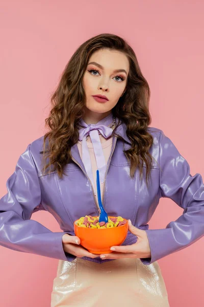 Conceptual, doll like, brunette woman with wavy hair pretending to be a doll, holding bowl with corn flakes and spoon, tasty breakfast, posing on pink background, looking at camera — Stock Photo