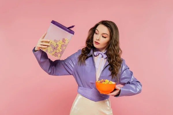 Konzeptfotografie, brünette Frau mit welligem Haar, die vorgibt, eine Puppe zu sein, Behälter mit Cornflakes in der Hand, leckeres Frühstück, posiert auf rosa Hintergrund, stylische lila Jacke — Stockfoto