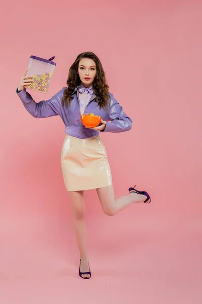 Концептуальная фотография, брюнетка с волнистыми волосами, притворяющаяся куклой, стоя на одной ноге, держа контейнер с кукурузными хлопьями, завтрак, позируя на розовом фоне, стильная фиолетовая куртка — стоковое фото
