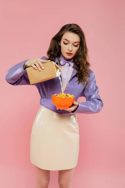 Concepto de fotografía, muñeca como, mujer joven con el pelo ondulado celebración bowl con hojuelas de maíz, verter la leche de la caja de cartón, delicioso desayuno, posando sobre fondo rosa, elegante chaqueta púrpura - foto de stock