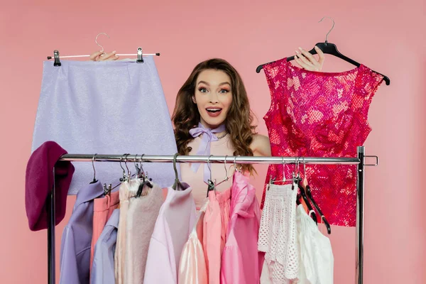 Consumismo, mujer joven sorprendida con el pelo ondulado morena de pie cerca de rack con ropa, mostrando ropa de moda, concepto de selección de armario, hermosa modelo mirando a la cámara en el fondo rosa - foto de stock