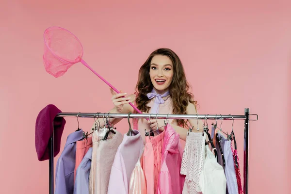 Consumerism, fashion photography, cheerful and young woman holding net on pink background, standing near rack with clothes, wardrobe selection, fashion and trends — Stock Photo