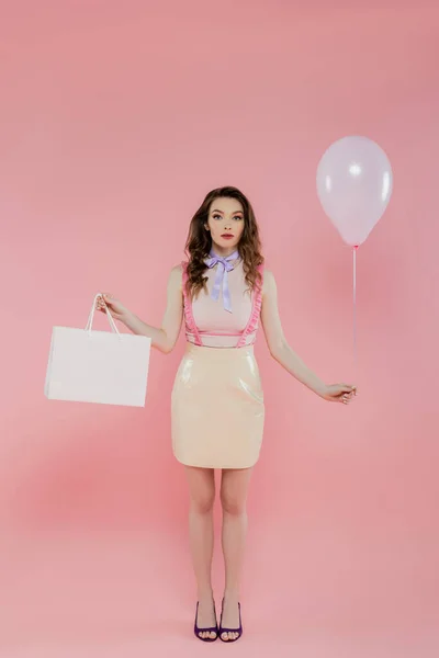 Модна фотографія, приваблива і молода жінка тримає повітряну кулю і сумку на рожевому фоні, позуючи як ляльку, стоїть і дивиться на камеру, одяг для дівчаток, споживач — стокове фото