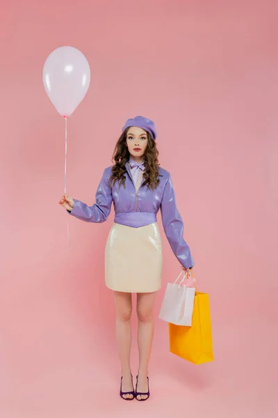Fotografia di moda, attraente e giovane donna che tiene palloncino e borse della spesa su sfondo rosa, in posa come una bambola, in piedi e guardando la fotocamera, alla moda, consumismo, vestito viola — Foto stock
