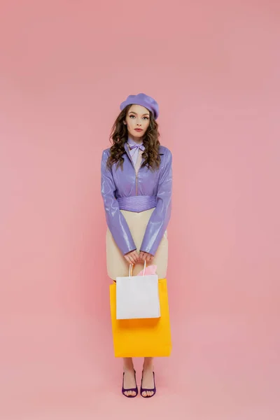 Consumerism, fashion photography, attractive young woman in beret holding shopping bags on pink background, posing like a doll, standing and looking at camera, trendy outfit, consumerism — Stock Photo