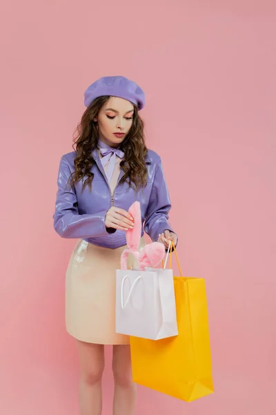 Консьюмеризм, модная фотография, привлекательная молодая женщина в берете, держащая сумки для покупок на розовом фоне, беря повязку для ушей кролика, консьюмеризм, стоя в модной куртке и юбке, — стоковое фото