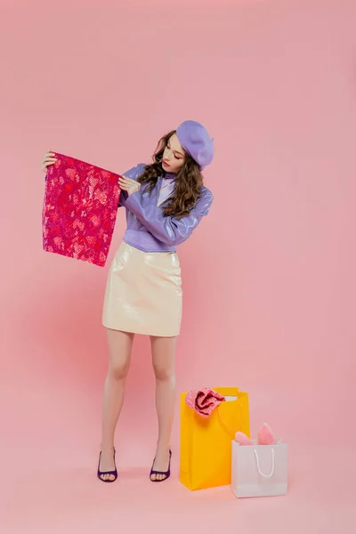 Mode et consumérisme, attrayant jeune femme en béret tenant des vêtements de couleur vibrante près des sacs à provisions sur fond rose, consumérisme, debout dans une veste et une jupe en cuir à la mode, pleine longueur — Photo de stock