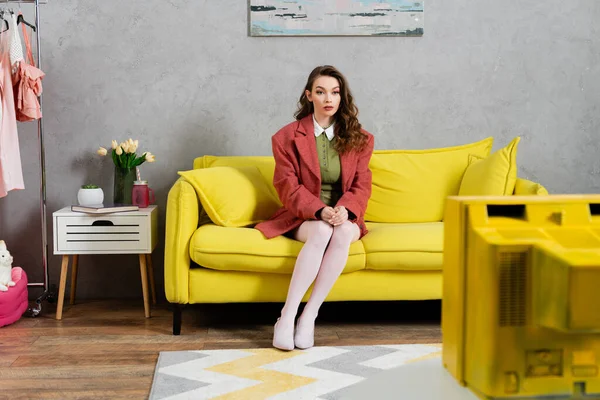 Concept photography, beautiful well dressed young woman with wavy hair sitting on yellow couch, stylish house interior, vase with tulips, watching tv, posing like a doll, housewife lifestyle — Stock Photo
