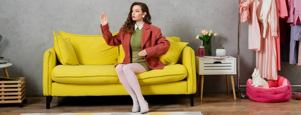 Fotografía conceptual, mujer actuando como una muñeca y sentada en un sofá amarillo, haciendo un gesto antinatural en la sala de estar moderna, interior de la casa moderna, juego de roles, vida de la muñeca, bien vestido, pancarta - foto de stock