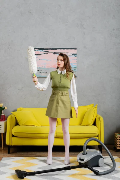 Housekeeping concept, young woman with wavy hair standing on clean carpet and holding dust brush near vacuum cleaner, housewife in dress, domestic life, posing like a doll — Stock Photo