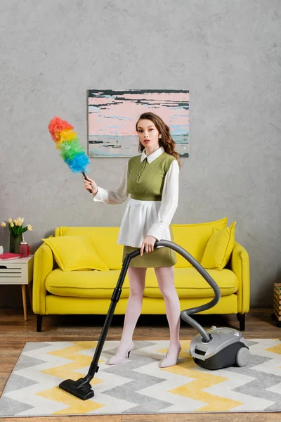 Housekeeping concept, young woman with wavy hair standing on carpet and holding dust brush, using vacuum cleaner, housewife in dress and white tights, domestic life, posing like a doll — Stock Photo