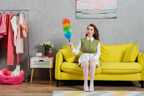 Concepto de limpieza, mujer joven con el pelo ondulado sentado en el sofá y la celebración de cepillo de polvo, ama de casa en vestido y medias blancas mirando a la cámara, la vida doméstica, posando como una muñeca - foto de stock