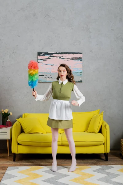Housekeeping concept, young woman with wavy hair standing and holding dust brush, housewife in dress and white tights looking at camera, gesturing unnaturally, domestic life, posing like a doll — Stock Photo