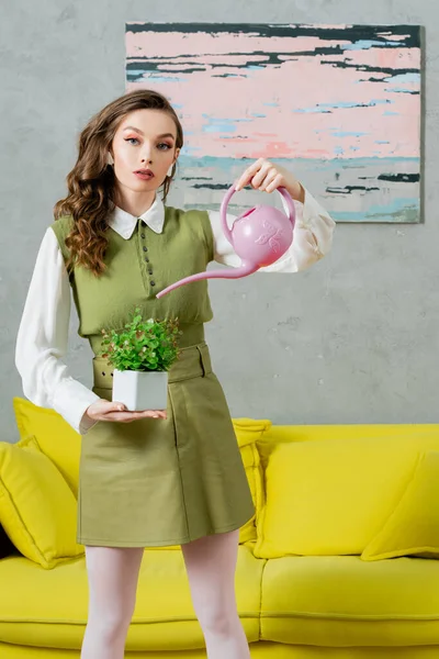 Eco-friendly, housekeeping concept, young woman with wavy hair watering green plant, growth and botany, housekeeping, beautiful housewife looking at camera and standing in living room — Stock Photo