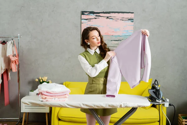 Концепція домогосподарства, молода жінка з хвилястим волоссям тримає чистий светр, домогосподарка виконує свої щоденні обов'язки, спосіб життя, домашні справи, прасування на день прання, домашні завдання — стокове фото