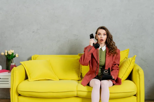 Phone call, attractive woman with wavy hair sitting on yellow couch, housewife talking on retro telephone, acting like a doll, looking at camera with opened mouth, shock, grey wall — Stock Photo