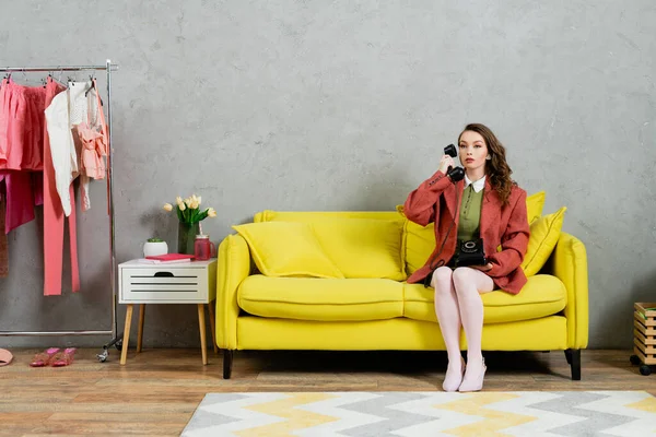 Coup de fil, jolie femme aux cheveux ondulés assise sur un canapé jaune, femme au foyer parlant au téléphone rétro, posant comme une poupée, regardant ailleurs, intérieur moderne, salon — Photo de stock