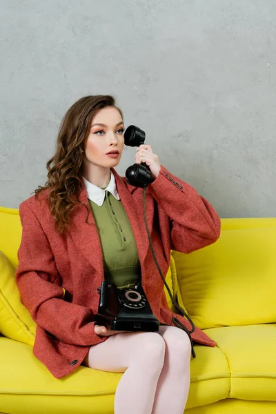 Vintage vibes, beautiful woman with brunette and wavy hair sitting on yellow couch, housewife talking on retro telephone, posing like a doll, looking away, modern interior, living room — Stock Photo