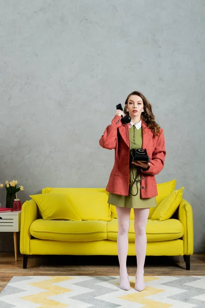 Vintage vibes, beautiful woman with brunette and wavy hair standing near yellow couch, housewife talking on retro telephone, posing like a doll, looking at camera, modern interior, living room — Stock Photo