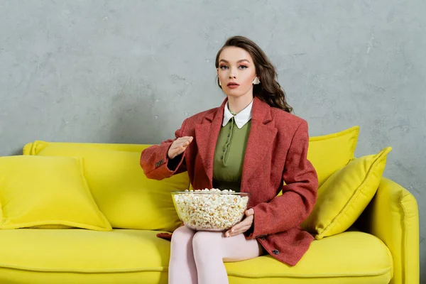 Pretty girl acting like a doll, concept photography, young woman with wavy holding bowl with popcorn, gesturing unnaturally, salty snack, home entertainment, sitting on comfortable yellow sofa — Stock Photo