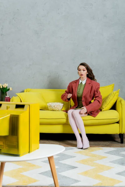 Mujer actuando como una muñeca, fotografía conceptual, modelo con cabello ondulado morena sentada en un sofá amarillo junto a un tazón de palomitas de maíz, viendo televisión, entretenimiento en el hogar, ocio, fotografía conceptual - foto de stock