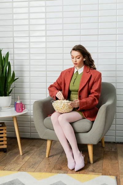 Концептуальна фотографія, молода жінка з брюнеткою хвилясте волосся, сидить на зручному кріслі, діє як лялька, дивлячись на миску з попкорном, дозвілля, домашні розваги, перекус фільму — стокове фото