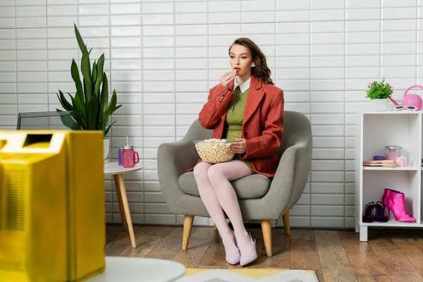 Konzeptfotografie, junge Frau mit brünetten welligen Haaren sitzt auf bequemen Sesseln, schaut fern und isst Popcorn, Freizeit, Home Entertainment, Filmsnack, puppenhaft — Stockfoto