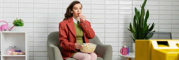 Concept photography, jeune femme avec des cheveux bruns ondulés assis sur un fauteuil confortable, regarder la télévision et manger du pop-corn, loisirs, divertissement à la maison, snack film, poupée comme, bannière — Photo de stock