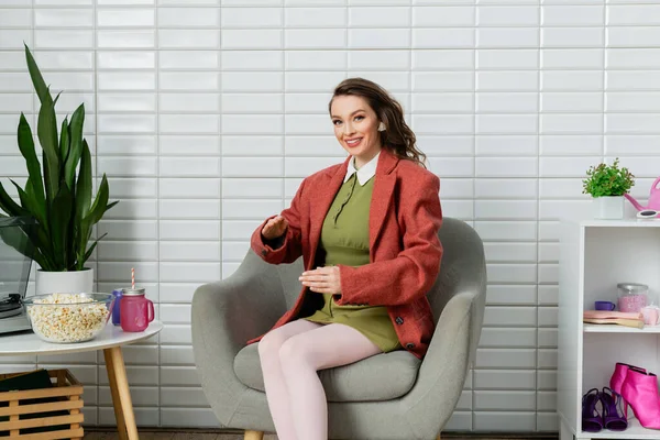 Концептуальная фотография, счастливая молодая женщина с волнистыми волосами, сидящая на удобном кресле рядом с миской с попкорном, действующая как кукла и неестественно жестикулирующая, кинозакуска, смотрящая в камеру — стоковое фото