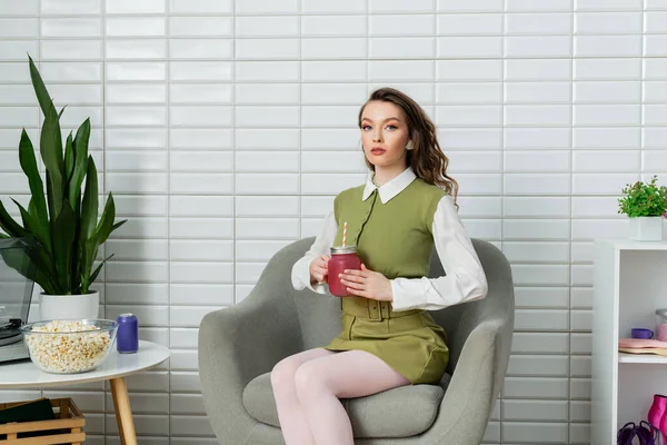Concepto de fotografía, mujer joven con cabello ondulado morena sentado en un sillón cómodo, actuando como una muñeca, sosteniendo bebida refrescante, tazón con palomitas de maíz, merienda de la película - foto de stock