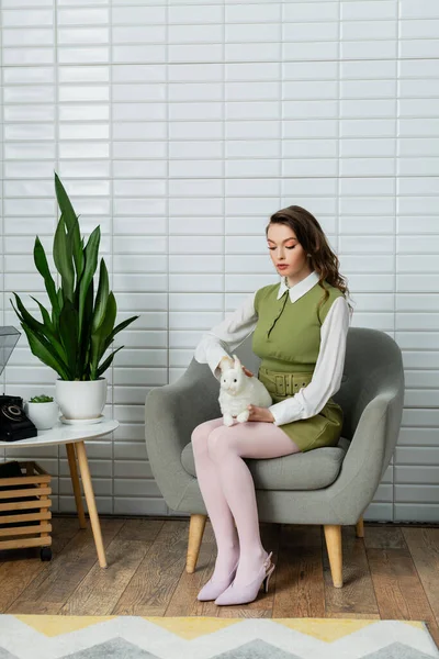 Femme agissant comme une poupée, belle femme assise sur un fauteuil gris confortable et tenant lapin jouet, plantes vertes et téléphone rétro sur la table, concept photographie — Photo de stock