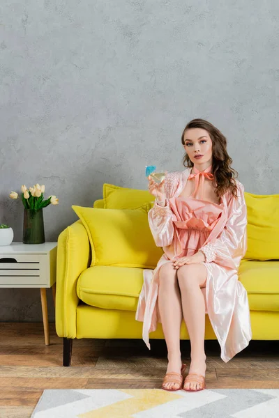Концептуальная фотография, женщина, действующая как кукла, домашняя жизнь, домохозяйка в розовом наряде с шелковым халатом, держащая коктейль в стекле, смотрящая на камеру и сидя на желтом автобусе в современной гостиной — стоковое фото