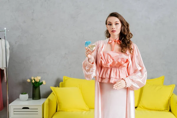 Концептуальная фотография, женщина, действующая как кукла, домашняя жизнь, домохозяйка в розовом наряде с шелковым халатом, держащая коктейль в стекле, жестикулируя и стоя рядом с желтым тренером в современной гостиной — стоковое фото