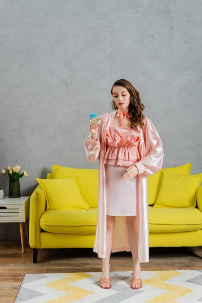 Концептуальна фотографія, молода жінка діє як лялька, домашнє життя, домогосподарка в рожевому вбранні з шовковим халатом, що тримає коктейль у склі, жестикулює і стоїть біля жовтого тренера в сучасній вітальні — стокове фото