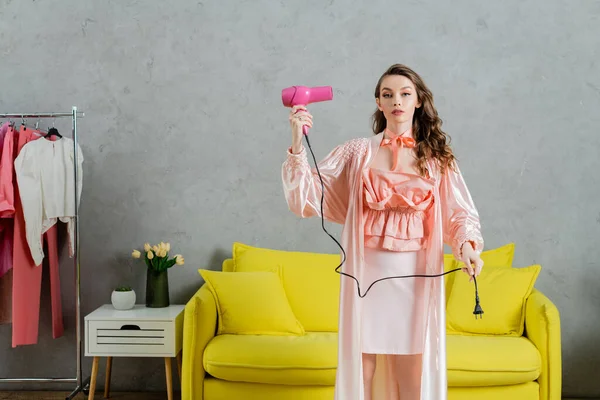 Концептуальна фотографія, жінка діє як лялька, домашнє життя, домогосподарка в рожевому шовковому халаті тримає фен і вилка, стоїть біля жовтого тренера в сучасній вітальні, прикидаючись сухим волоссям — стокове фото