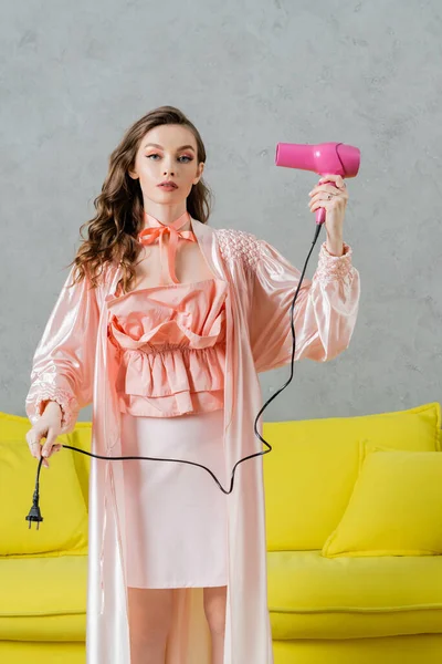 Концептуальная фотография, женщина, действующая как кукла, красивая домохозяйка в розовом шелковом халате, держащая фен и вилку, стоящая рядом с желтым тренером в современной гостиной, притворяющаяся сушкой волос — стоковое фото