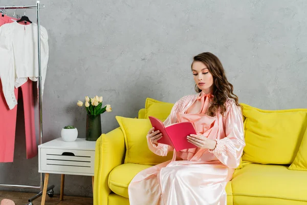 Konzeptfotografie, Frau mit brünetten welligen Haaren, häusliches Leben, attraktive Hausfrau liest Buch, sitzt auf gelbem Sofa, komfortables Wohnen, häuslicher Lebensstil — Stockfoto