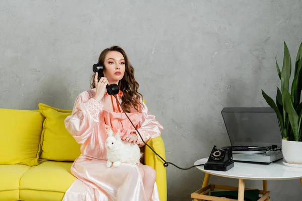 Vintage vibes, beautiful woman with brunette and wavy hair sitting on yellow couch, housewife talking on retro telephone, posing like a doll, looking away, vinyl record player — Stock Photo