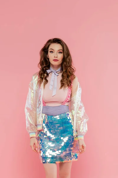 Beautiful woman with wavy hair posing like a doll on pink background, conceptual photography, girly outfit, model in skirt with sequins and transparent jacket looking at camera — Stock Photo