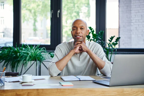 Ptosis syndrome, bold african american businessman with eye syndrome looking at camera near laptop on desk, myasthenia gravis, cheerful dark skinned office worker, diversity and inclusion — Stock Photo