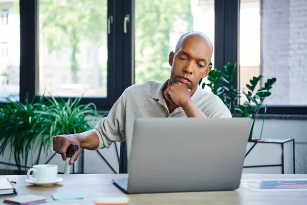 Ptosis syndrome, bold african american businessman using laptop, dark skinned office worker with myasthenia gravis disease holding walking cane, diversity and inclusion, corporate culture — Stock Photo