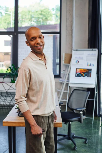 Bold dark skinned man with myasthenia gravis disease standing with walking cane, cheerful african american office worker with ptosis eye syndrome, inclusion, infographics and flipchart on background — Stock Photo