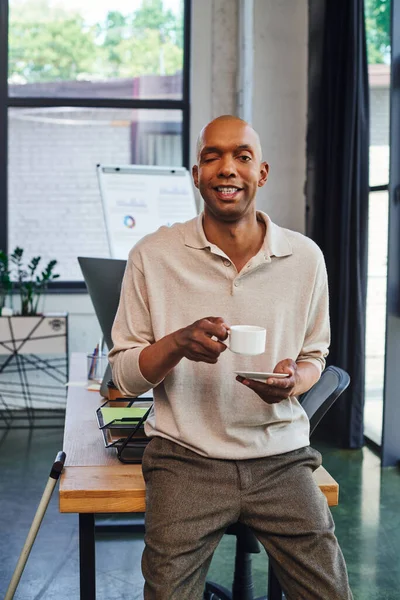 Homme à la peau foncée avec la maladie de la myasthénie grave debout avec une tasse de café près de la canne à pied, travailleur de bureau afro-américain joyeux avec le syndrome de l'oeil de ptose, inclusion, graphiques sur le fond — Photo de stock