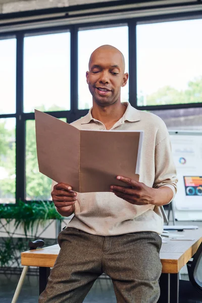 Happy dark skinned man with myasthenia gravis disease holding folder and standing near desk and walking cane, bold african american office worker with ptosis eye syndrome, inclusion, work — Stock Photo