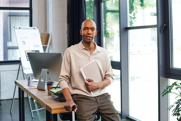 Homme à la peau foncée avec la maladie de Myasthénie Gravis tenant dossier et debout avec la canne à pied, travailleur de bureau afro-américain audacieux avec le syndrome de l'oeil de ptose, inclusion, moniteur et graphiques — Photo de stock