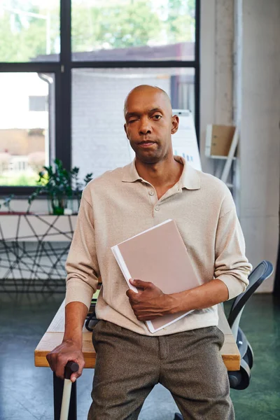 Dark skinned man with myasthenia gravis disease holding folder and standing with walking cane, bold african american office worker looking at camera, inclusion, corporate culture — Stock Photo
