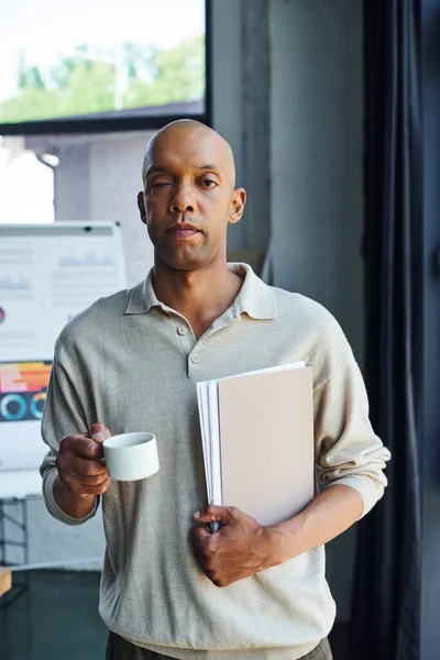 African american man with myasthenia gravis disease holding folder and cup of coffee, bold and dark skinned office worker with ptosis eye syndrome, inclusion, professional headshots — Stock Photo